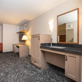 Accessible hotel room with a large desk, open vanity with mirror, armchair, and patterned carpet. Neutral colors and modern furnishings.
