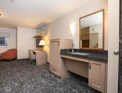 Accessible hotel room with a large desk, open vanity with mirror, armchair, and patterned carpet. Neutral colors and modern furnishings.