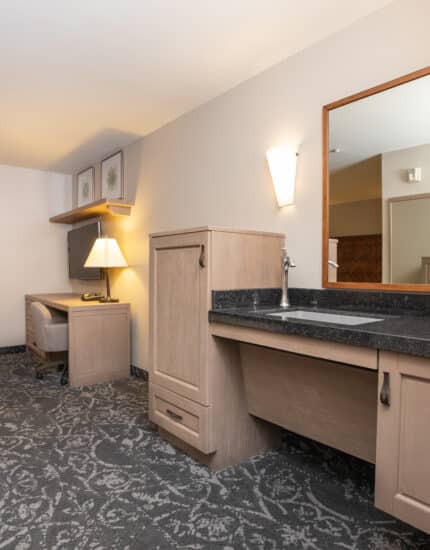 Accessible hotel room with a large desk, open vanity with mirror, armchair, and patterned carpet. Neutral colors and modern furnishings.