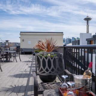 Rooftop patio with tables, chairs, and potted plants overlooking a cityscape with the Space Needle visible in the background.