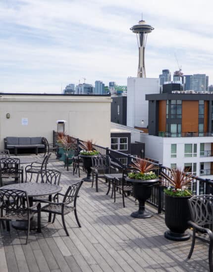 Rooftop patio with tables, chairs, potted plants, and a view of the Seattle skyline, including the Space Needle in the background.