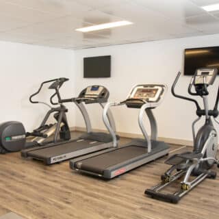 A gym room with two treadmills, an elliptical machine, a stationary bike, wall-mounted TV screens, and light wood flooring.