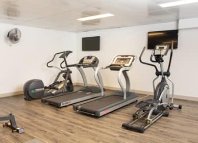 A gym room with two treadmills, an elliptical machine, a stationary bike, wall-mounted TV screens, and light wood flooring.