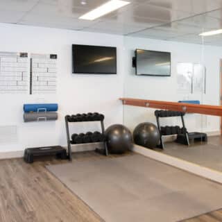 A gym room with free weights, two exercise balls, workout charts, two TVs, a mirror, and wooden floors. Yoga mats and a step platform are stored on the wall.