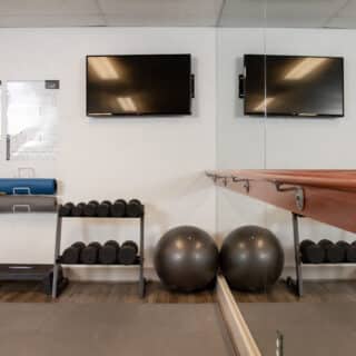 A fitness studio with exercise mats, dumbbells, stability balls, a step platform, two wall-mounted TVs, and a barre along a mirrored wall.