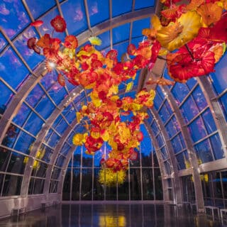 A glass-walled atrium features a large, colorful glass sculpture resembling flowers, suspended from the ceiling, illuminated against the evening sky.