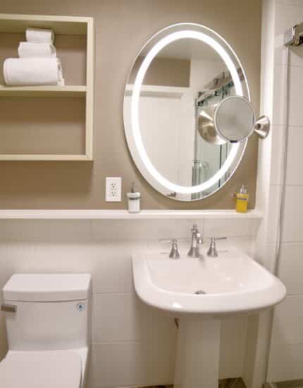 Modern bathroom with a round backlit mirror, white sink, toilet, wall shelves with towels, and a glass shower with mounted toiletries.