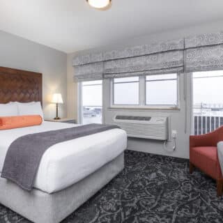 A hotel room with a neatly made bed, wooden headboard, orange bolster pillow, bedside lamps, red chair, desk, and large windows with patterned valances.
