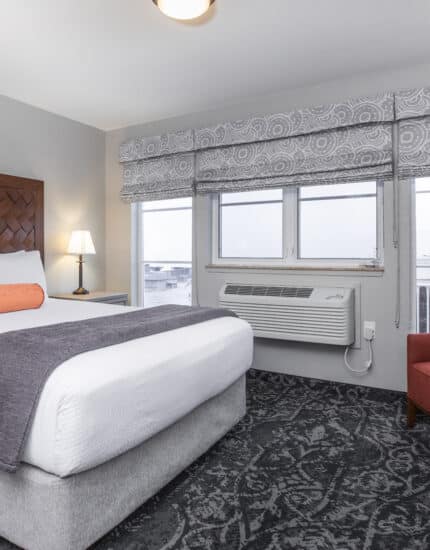 A hotel room with a neatly made bed, wooden headboard, orange bolster pillow, bedside lamps, red chair, desk, and large windows with patterned valances.