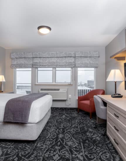 A hotel room with a bed, two desks with lamps, a wall-mounted TV, a red armchair, patterned carpet, and three windows with city views.