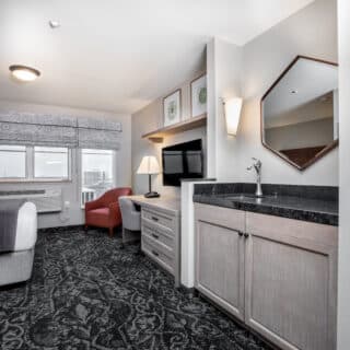 Modern hotel room with patterned carpet, a seating area by the window, a desk with a lamp, a wall-mounted TV, and a small kitchenette with a sink and hexagonal mirror.