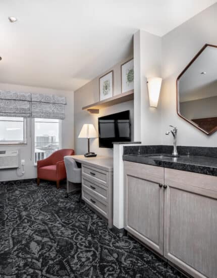Modern hotel room with a patterned carpet, seating area, desk, TV, lamp, window with city view, and a kitchenette with a sink and mirror.