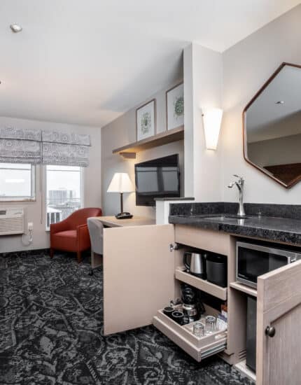 A hotel room with a sitting area, flat-screen TV, mini-fridge, microwave, coffee maker, and large window with city view.