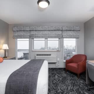 A hotel room with a bed, desk, two lamps, a red chair, and large windows showing a city view.
