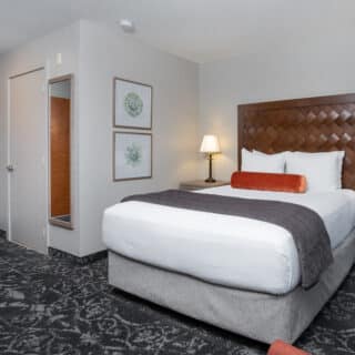 A hotel room with a queen bed, two nightstands with lamps, framed art on the wall, a mirror, and a door in the background.