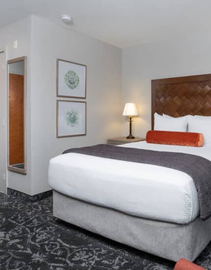 A hotel room with a queen bed, two nightstands with lamps, framed art on the wall, a mirror, and a door in the background.