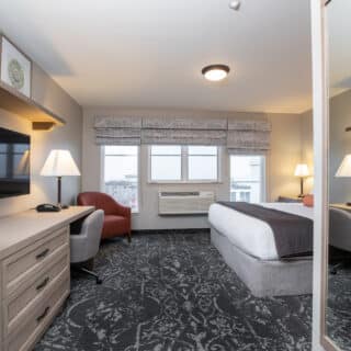 Hotel room with a bed, desk and chair, flat-screen TV, armchair, lamps, framed art, large window, and patterned carpet. Mirror reflects part of the room.
