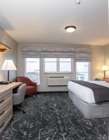 Hotel room with a bed, desk and chair, flat-screen TV, armchair, lamps, framed art, large window, and patterned carpet. Mirror reflects part of the room.