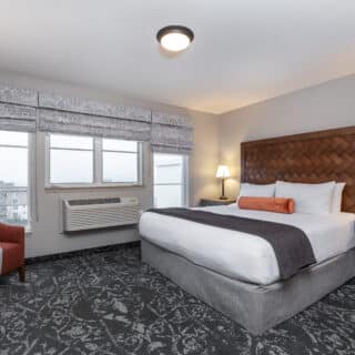 A hotel room with a king-size bed, two bedside tables with lamps, a desk and chair, an armchair, patterned carpet, and two windows with patterned valances.