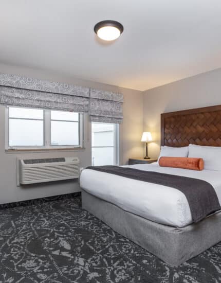 A hotel room with a king-size bed, two bedside tables with lamps, a desk and chair, an armchair, patterned carpet, and two windows with patterned valances.