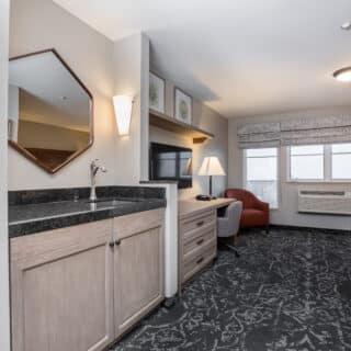 Hotel room with a kitchenette, TV, desk, armchair, bed, windows with patterned curtains, and neutral-colored furnishings.
