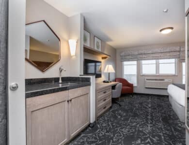Hotel room with a kitchenette, TV, desk, armchair, bed, windows with patterned curtains, and neutral-colored furnishings.