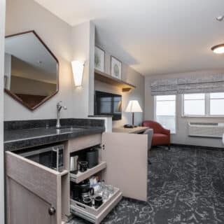 Hotel room with a queen bed, armchair, TV, desk, and open kitchenette cabinet displaying coffee and drink supplies. Windows let in natural light.