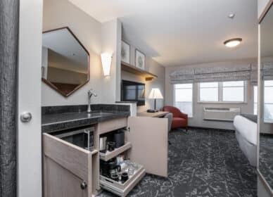 Hotel room with a queen bed, armchair, TV, desk, and open kitchenette cabinet displaying coffee and drink supplies. Windows let in natural light.