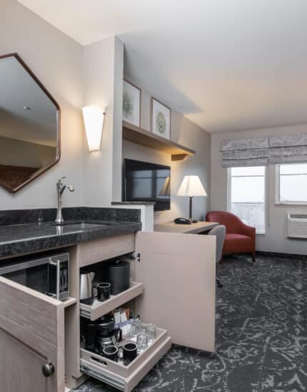Hotel room with a queen bed, armchair, TV, desk, and open kitchenette cabinet displaying coffee and drink supplies. Windows let in natural light.