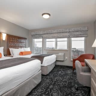 Hotel room with two beds, a desk with a chair and lamp, a TV, an armchair, and large window with city view. The room features neutral colors and patterned carpet.