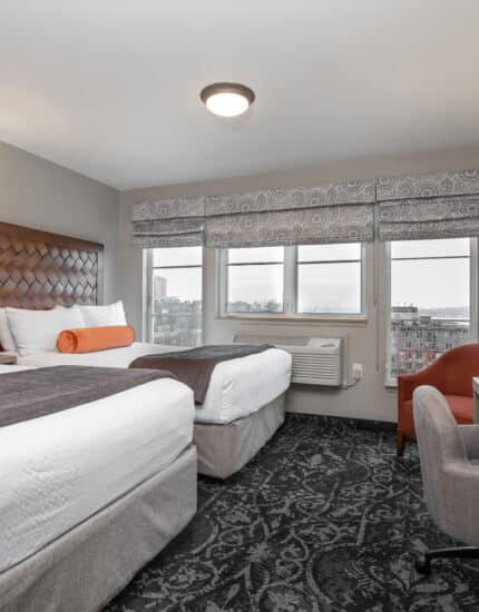 Hotel room with two beds, a desk with a chair and lamp, a TV, an armchair, and large window with city view. The room features neutral colors and patterned carpet.