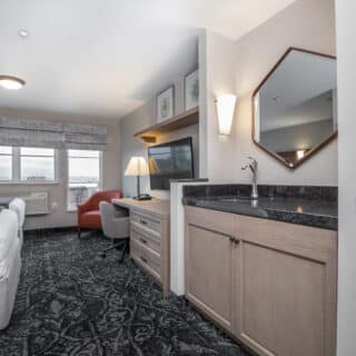 Hotel room with a bed, desk, TV, armchair, and a sink area with a mirror. Large windows provide natural light, and patterned carpet covers the floor.