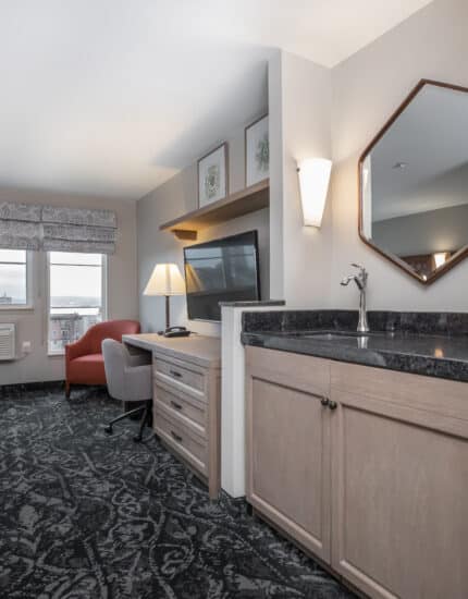 Hotel room with a bed, desk, TV, armchair, and a sink area with a mirror. Large windows provide natural light, and patterned carpet covers the floor.