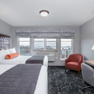 Hotel room with two double beds, a desk with a lamp and TV, an armchair, and large windows with a view outside.