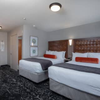 Hotel room with two double beds, dark headboards, orange bolster pillows, a desk, wall art, and a door leading to the hallway.