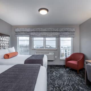 Modern hotel room with two double beds, a red armchair, work desk, lamp, and large windows showing a city and water view.