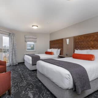 Hotel room with two queen beds, orange bolsters, gray throws, patterned carpet, two red chairs, a small table, and a door leading to a balcony.