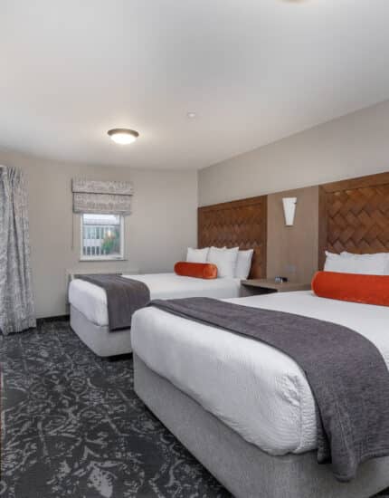 Hotel room with two queen beds, orange bolsters, gray throws, patterned carpet, two red chairs, a small table, and a door leading to a balcony.