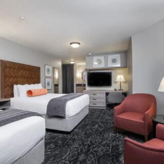 Hotel room with two double beds, a wall-mounted TV, a desk, two red armchairs, a lamp, and decorative artwork on the walls.