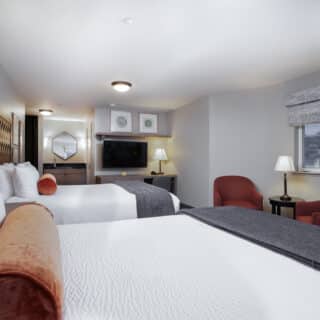 Hotel room with two double beds, a TV, a desk, two red chairs by a window, white bedding, and modern decor.