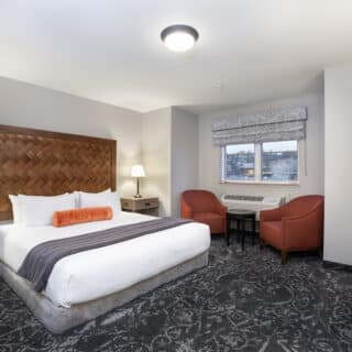 A hotel room with a large bed, two bedside tables with lamps, two red chairs by a window, a small table, and a desk with a chair.