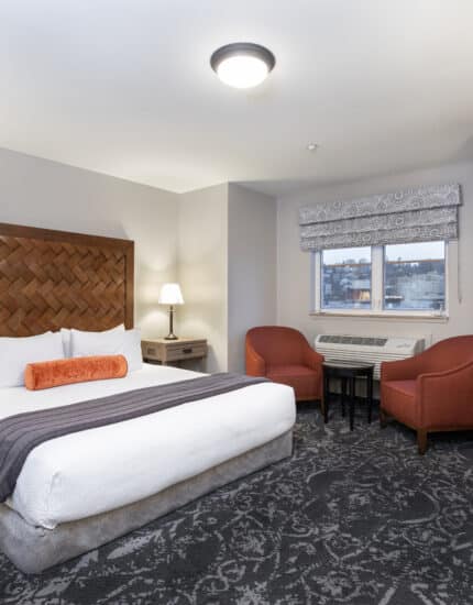 A hotel room with a large bed, two bedside tables with lamps, two red chairs by a window, a small table, and a desk with a chair.