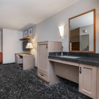 Hotel room with desk, chair, and lamp in the background; foreground features a sink with a mirror and cabinets, all in neutral tones.