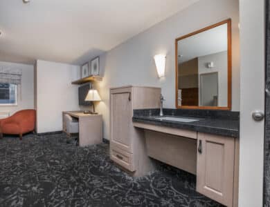 Hotel room with desk, chair, and lamp in the background; foreground features a sink with a mirror and cabinets, all in neutral tones.