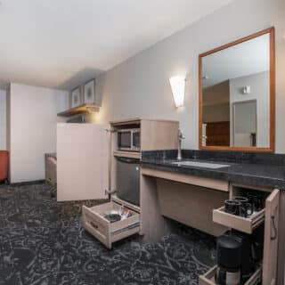 Hotel room kitchenette with open cabinets, a microwave, mini fridge, sink, coffee maker, mugs, and drawers. A window, chair, and table are visible in the background.