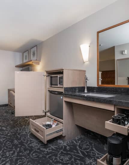 Hotel room kitchenette with open cabinets, a microwave, mini fridge, sink, coffee maker, mugs, and drawers. A window, chair, and table are visible in the background.