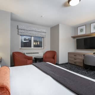 Hotel room with a bed, two red chairs by a window, a desk with a TV and chair, lamps on both sides, and framed artwork above the desk.