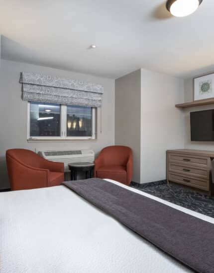 Hotel room with a bed, two red chairs by a window, a desk with a TV and chair, lamps on both sides, and framed artwork above the desk.