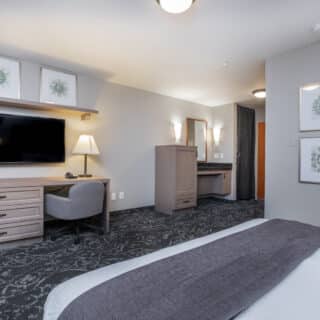 Modern hotel room with a bed, desk and chair, flat-screen TV, lamp, and wall art, featuring neutral tones and patterned carpet.