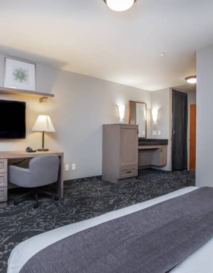 Modern hotel room with a bed, desk and chair, flat-screen TV, lamp, and wall art, featuring neutral tones and patterned carpet.